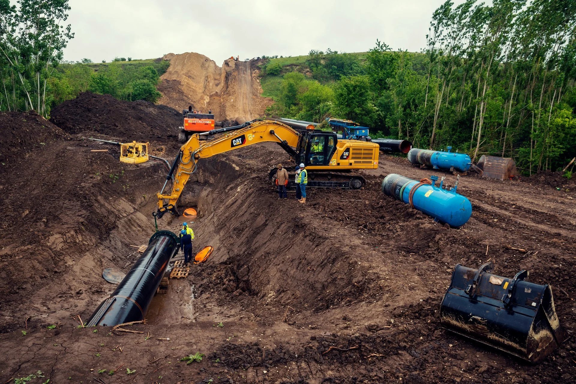 Heavy construction equipment including yellow excavator and black compactor working at an active excavation site with pipes and piled earth